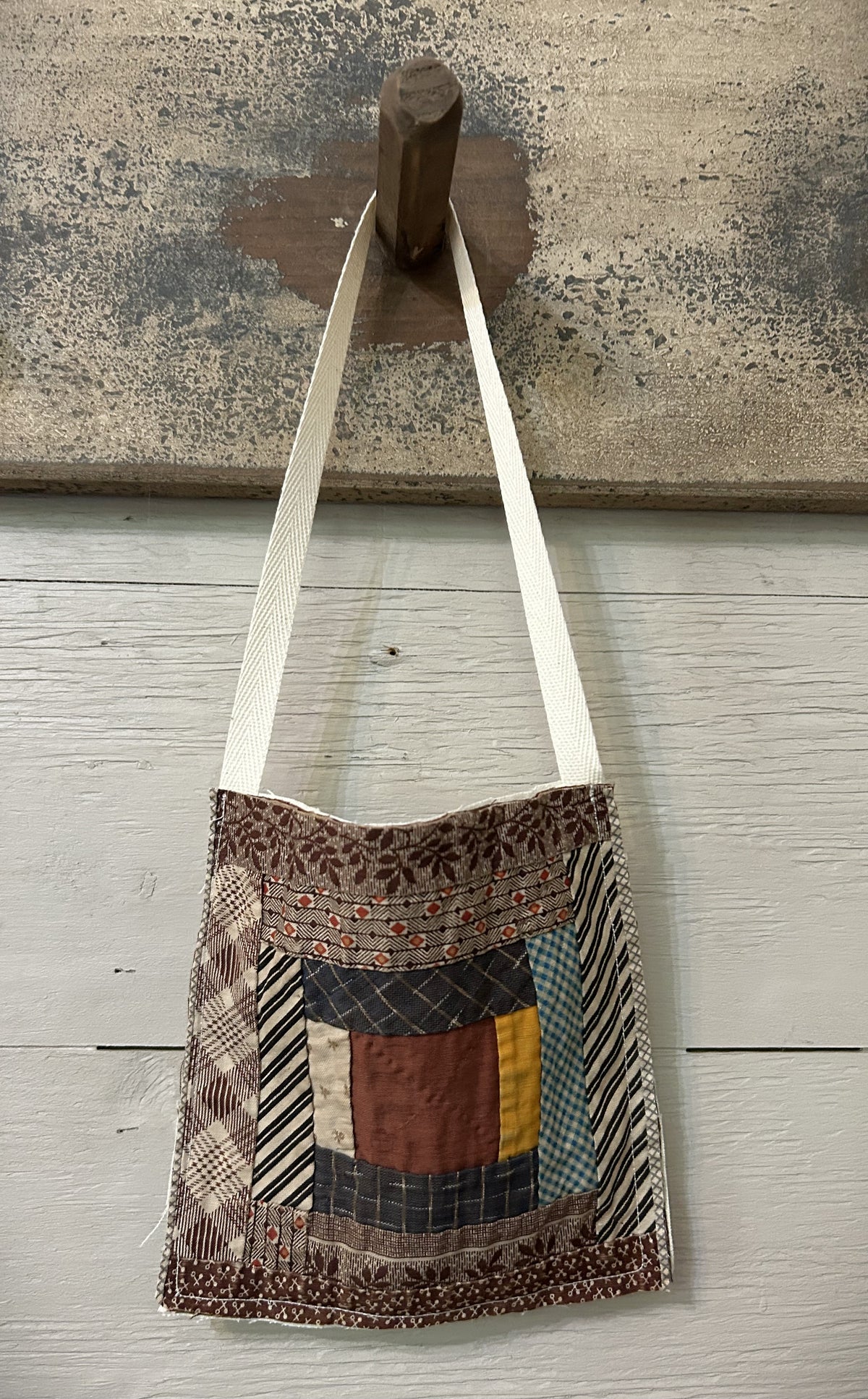 Primitive antique log cabin quilt block peg hanger in earth tones of brown, black and white stripe, blue-gray plaid, mustard gold and rust, hanging from natural cotton ribbon on a wooden peg