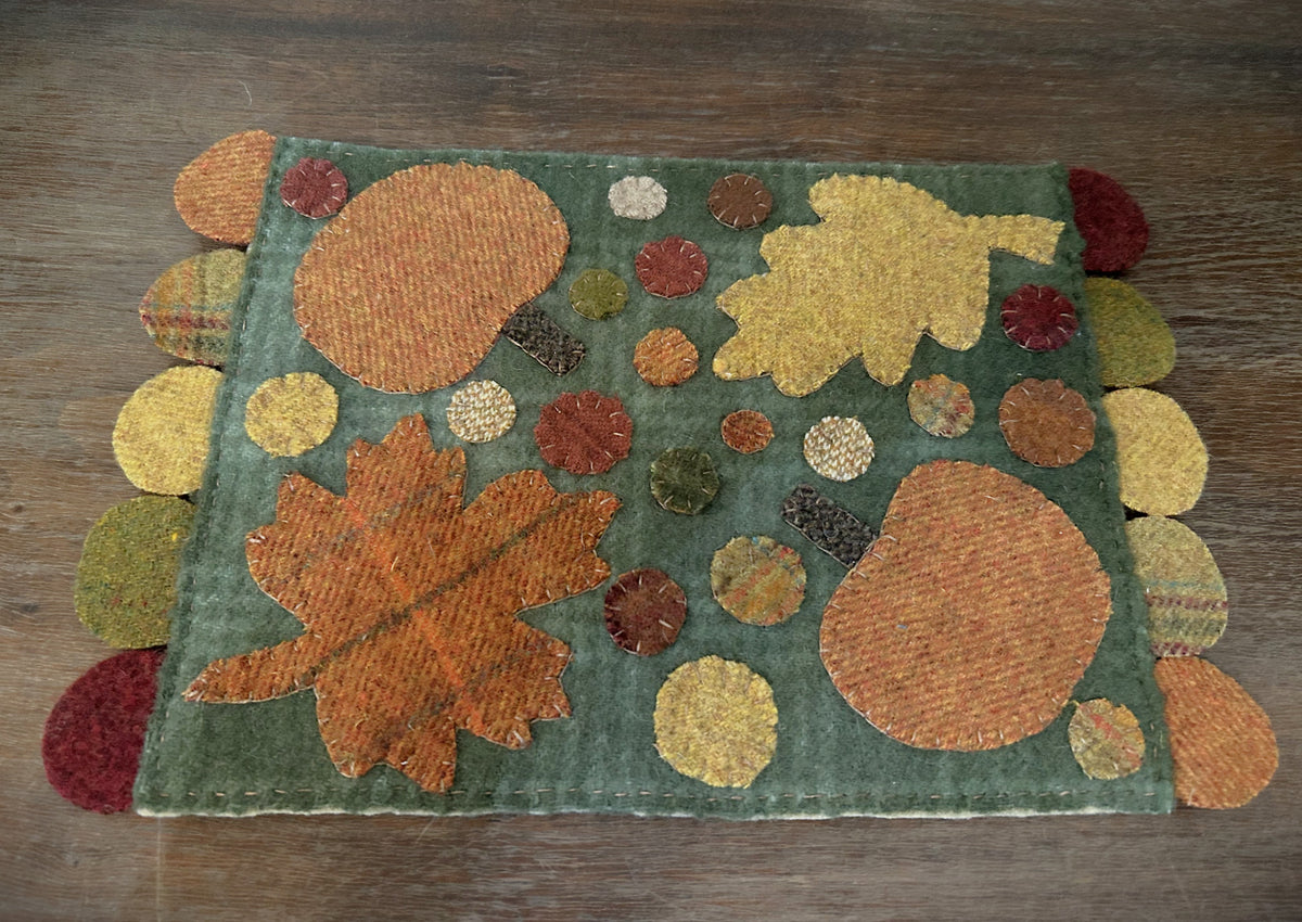 Handstitched primitive wool penny mat with green plaid background, burnt orange plaid and mustard leaf appliqués, pumpkins, and multi-colored penny circle edging in fall shades